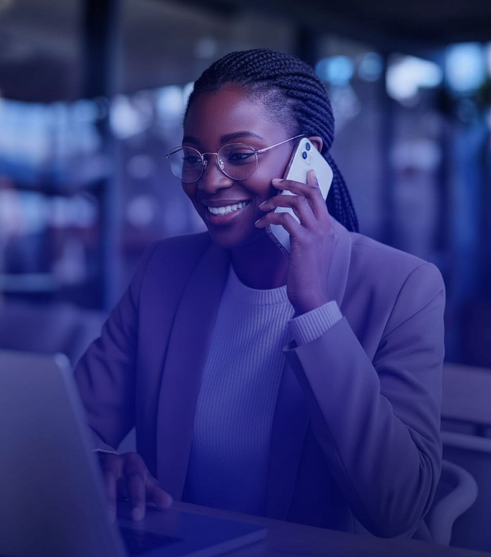 A professional African woman with braided hair and glasses, smiling while talking on her smartphone and working on a laptop in a modern office with a blue color overlay.